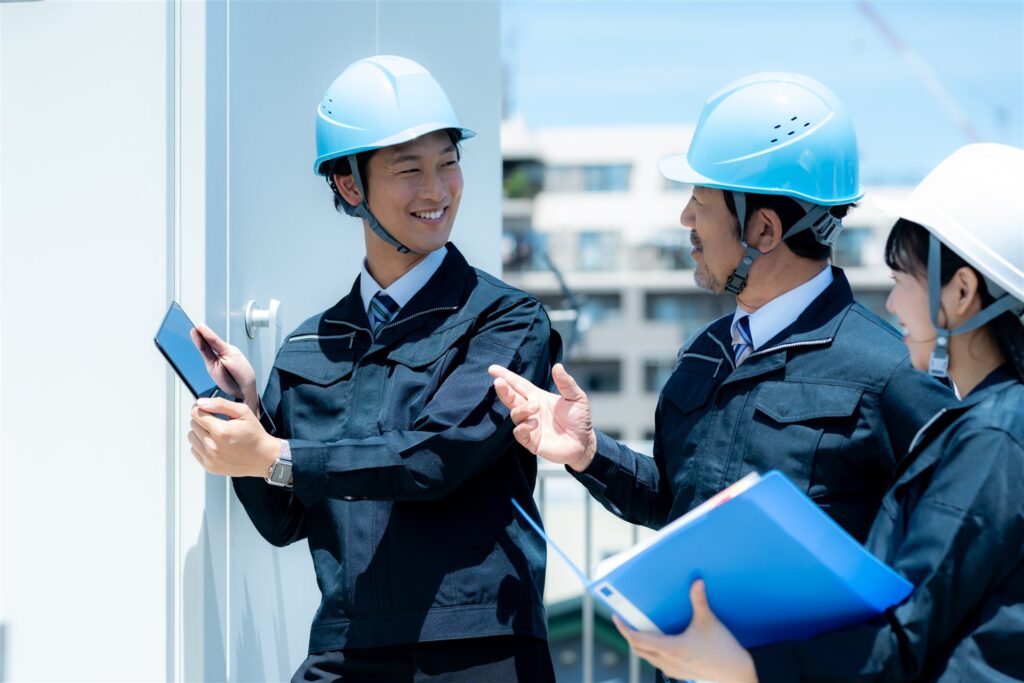 屋上でタブレットと書類を手に話す作業服の3人