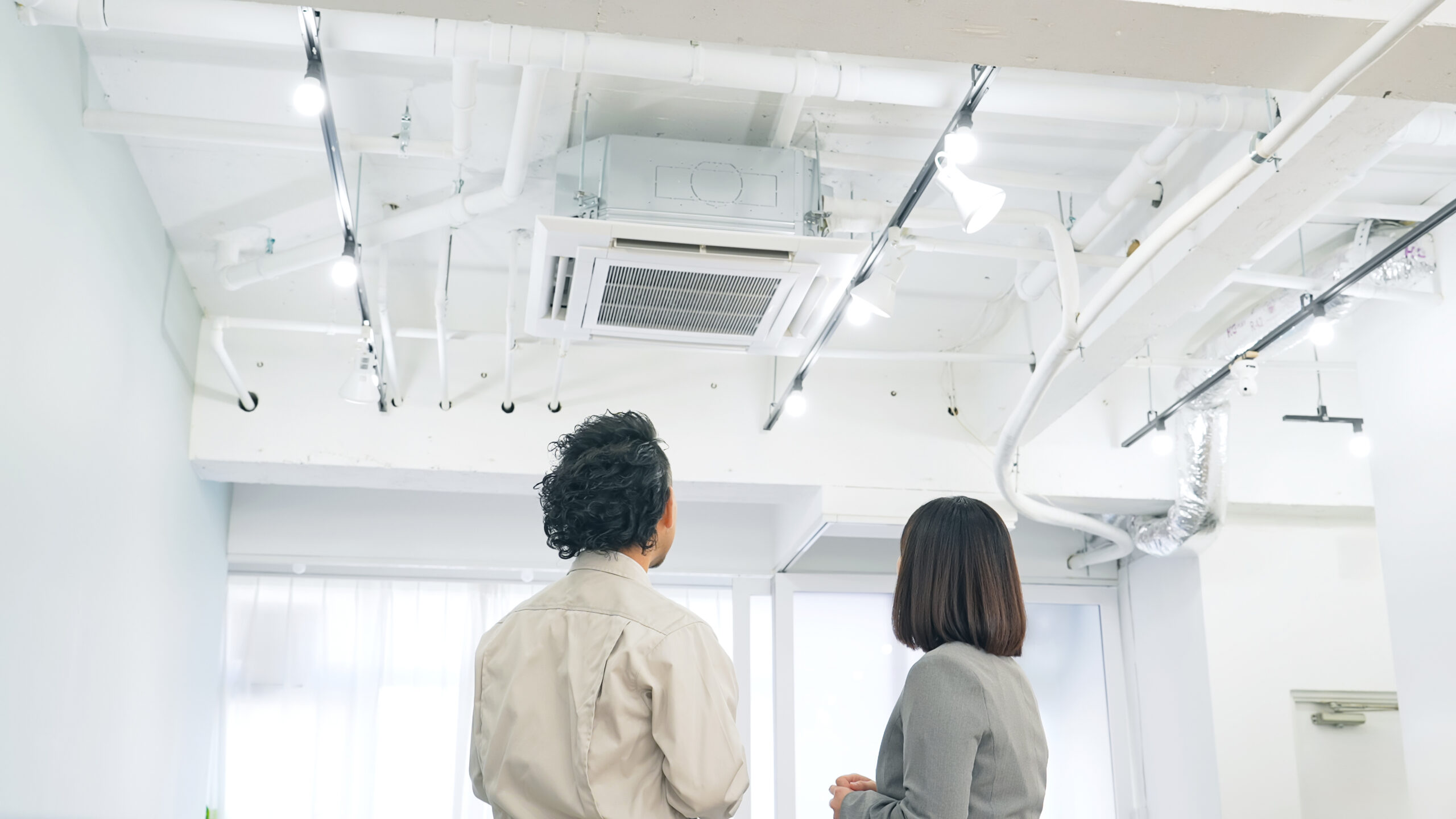 天井の空調と配管を見上げる作業着の人と同行者