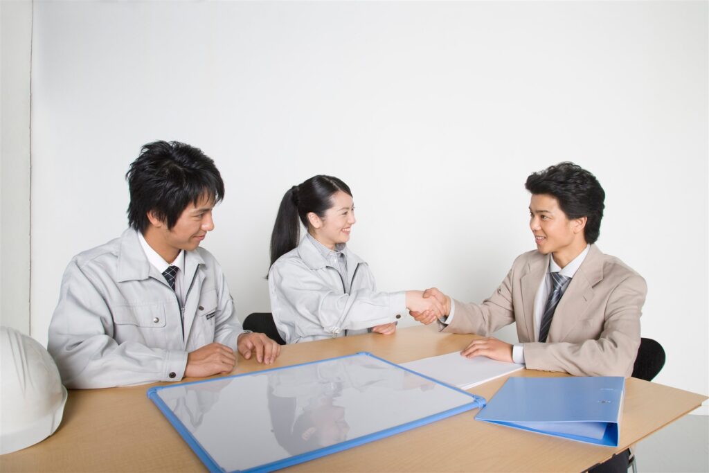 机をはさんで握手する作業着の2人と背広の男性