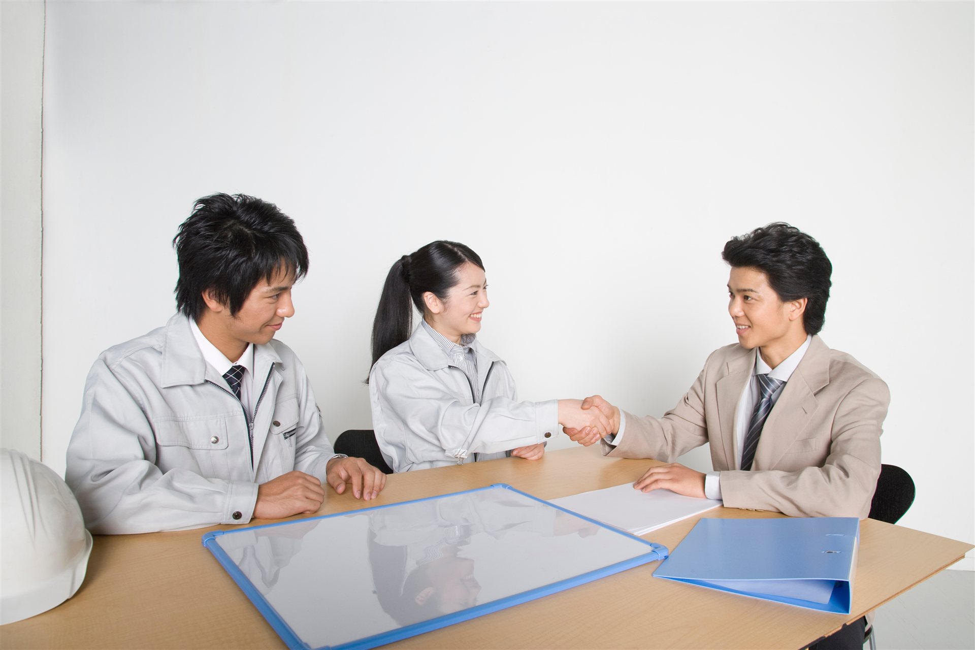 机をはさんで握手する作業着の2人と背広の男性