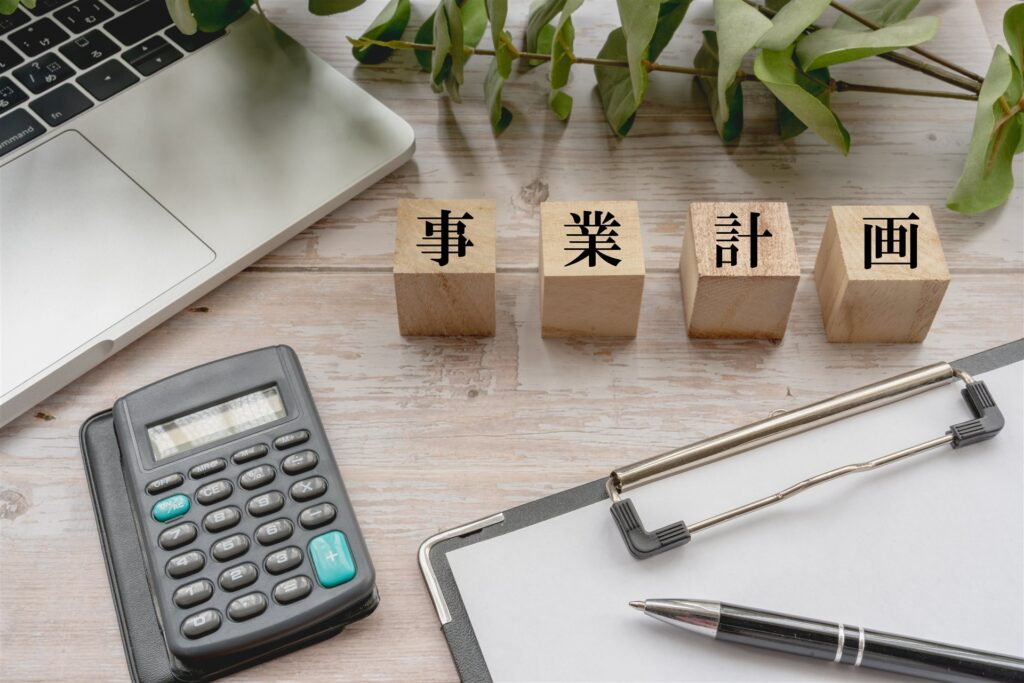 「事業計画」の木の札と電卓とノートパソコン