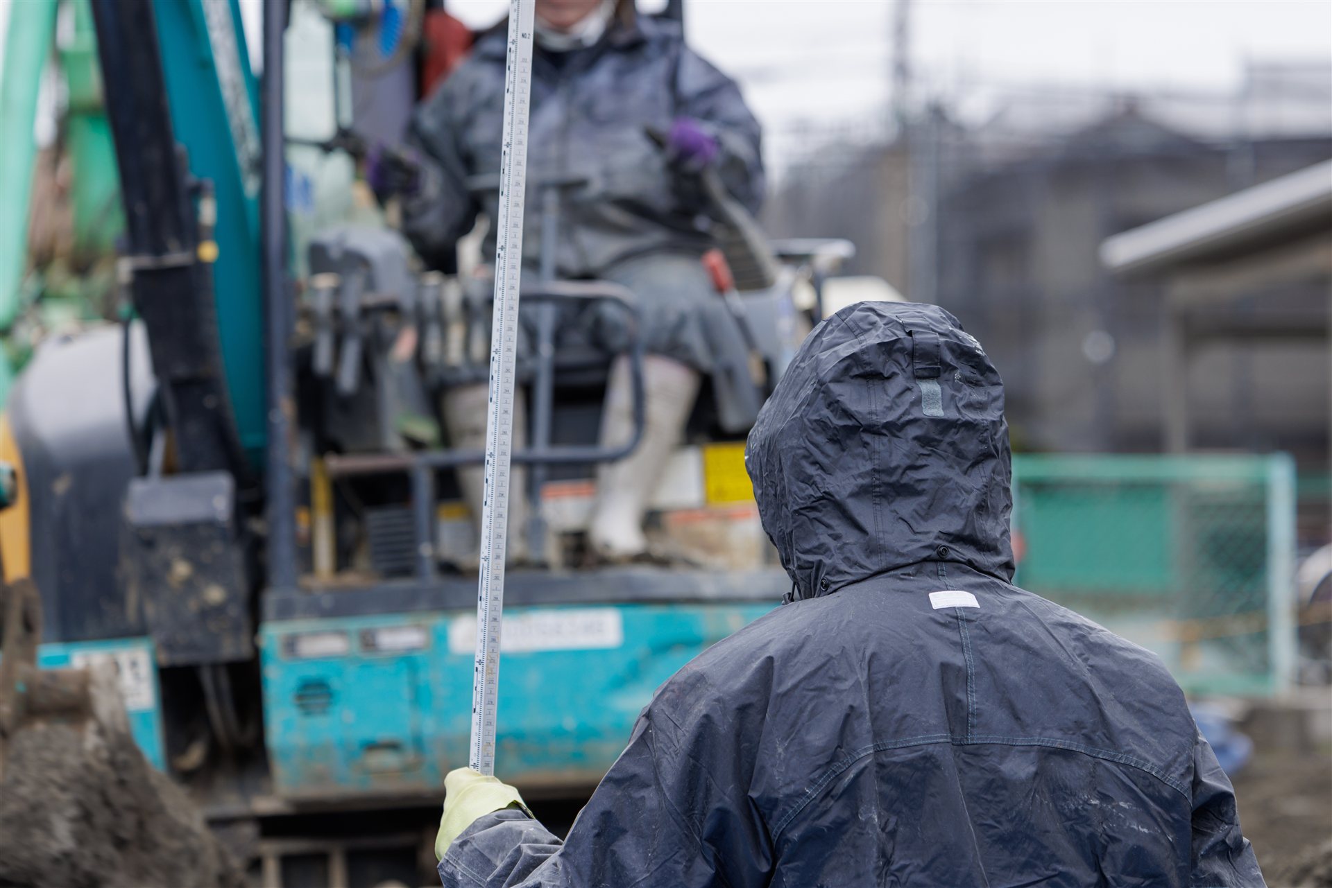 雨の中で雨がっぱを着て作業する人と重機(後ろ姿)