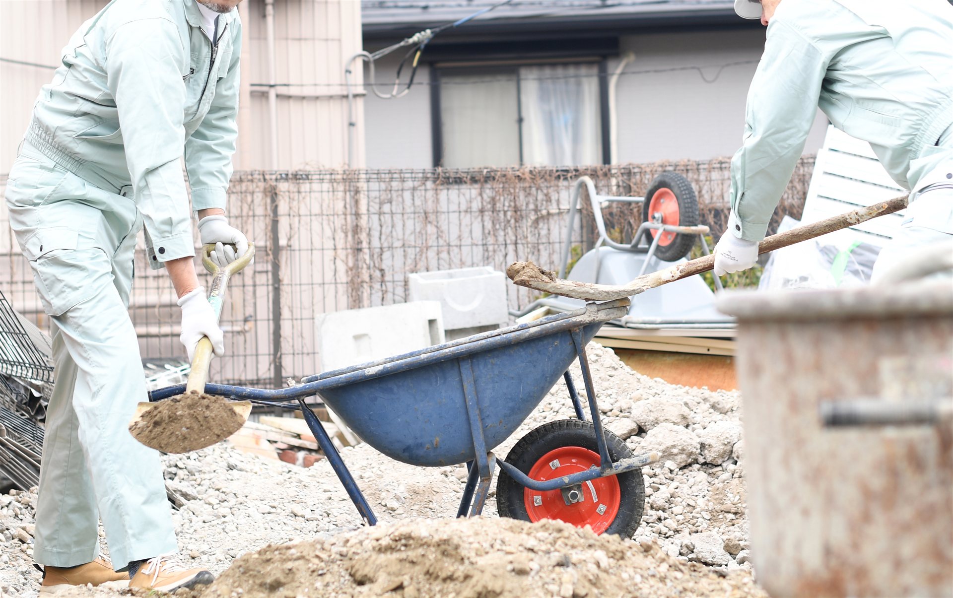 スコップで土を一輪車に入れる作業着の2人