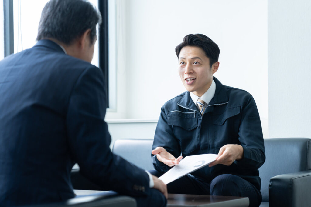 作業服の男性が書類を手に向かいの人へ説明する