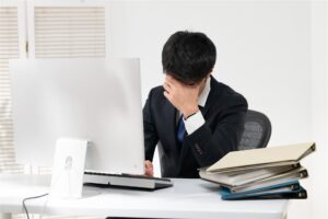 書類の束の横で頭を抱えるスーツの男性