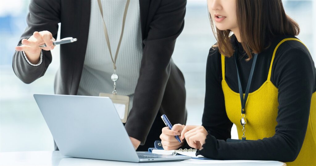 ノートパソコンを指しながらメモを取る人に話す社員