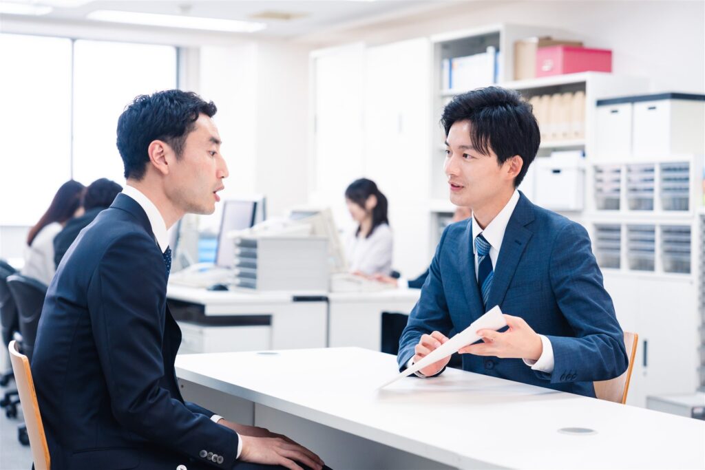 事務所の机で書類を手に話すスーツの男性2人